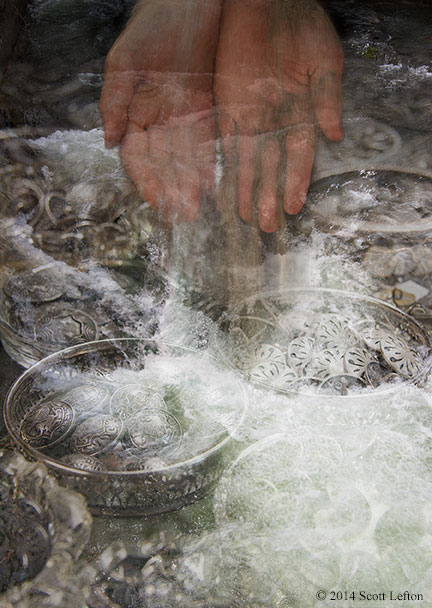 Water cascades over bowls of ornale metal objects, as a pair of semi-transparent hands pour water and sand into the flood.