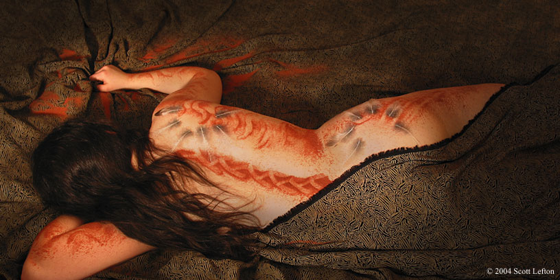 A woman lies in bed, clutching the sheets.  She's covered in feather designs and red sand painting.